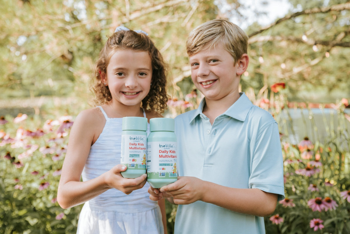 Two smiling kids in a garden, each holding a bottle of True Littles® Daily Kids Multivitamin