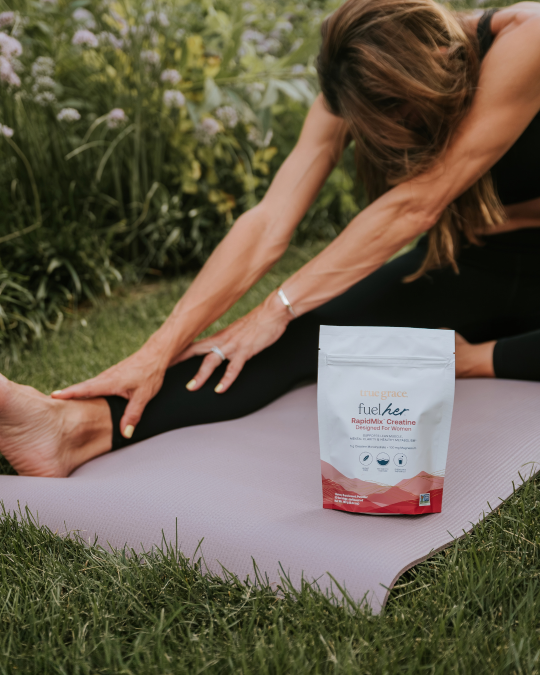 A woman in black activewear stretches on a pink yoga mat outdoors in a garden, with a white and red bag of FuelHer RapidMix Creatine supplement placed on the mat.