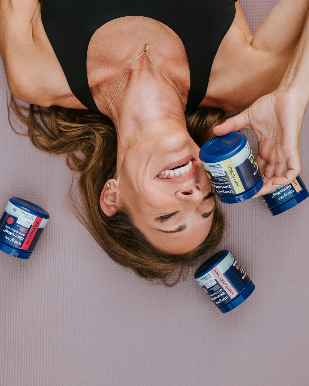 A woman lying on a pink yoga mat holds three blue supplement jars while two more jars rest on the mat around her. She is wearing a black sports bra and a delicate gold necklace.