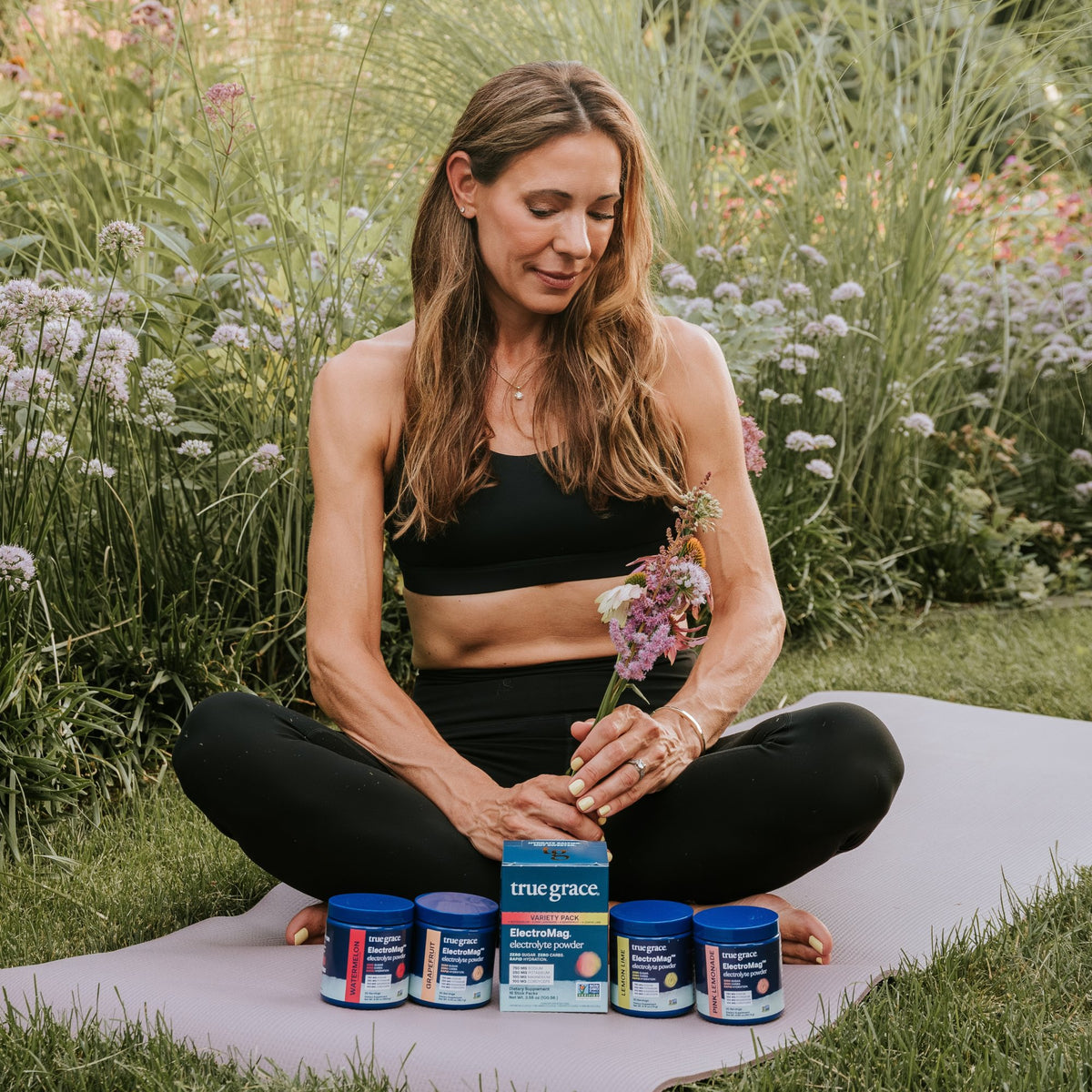 A woman sitting on a yoga mat in a garden with an ElectroMag® Variety Pack box and four canisters