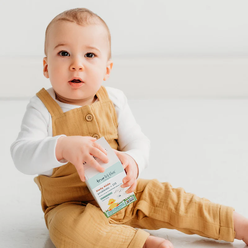 Baby in mustard overalls holding a box of True Littles Daily Kids Probiotics