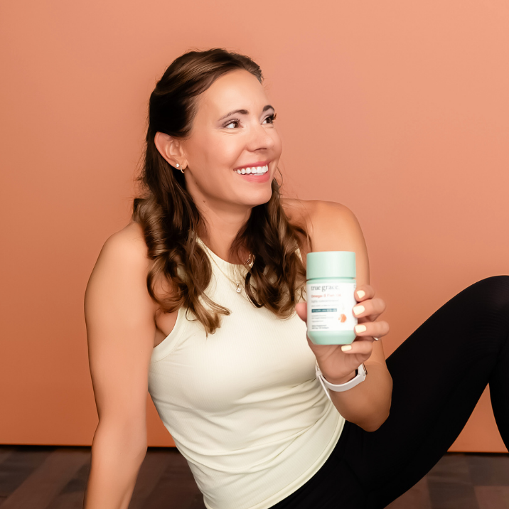 Woman sitting on a yoga mat holding a supplement container against a peach-colored wall.