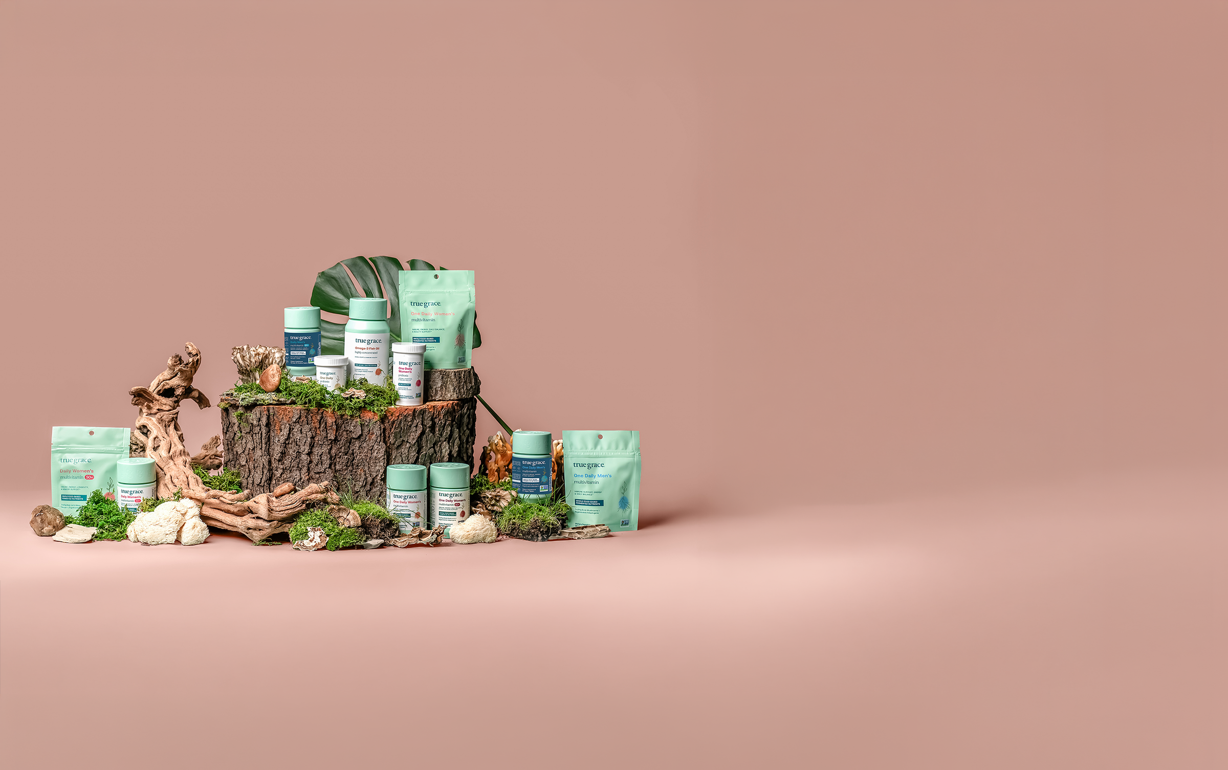 A styled product display of multiple True Grace supplement and wellness items including pouches, and a box, arranged on a rustic tree stump with moss, driftwood, and small plants against a solid dusty pink background.