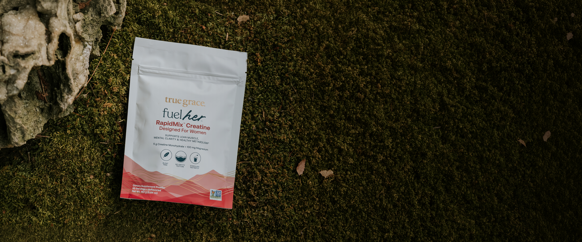 A pouch of FuelHer RapidMix Creatine powder sits on a bed of dark green moss next to a piece of weathered driftwood.