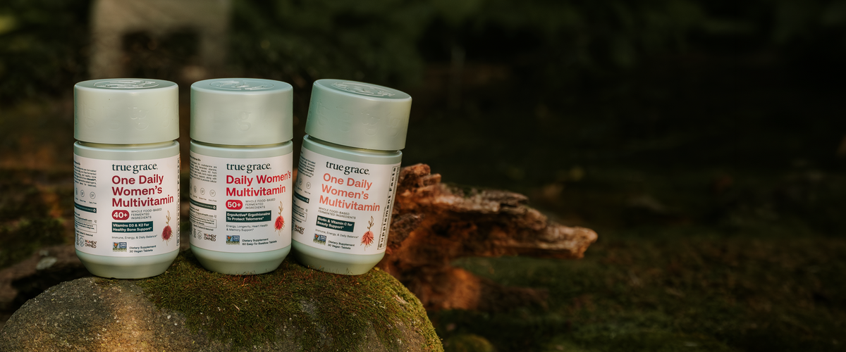 Three white bottles of true grace One Daily Women's Multivitamin arranged on a mossy log outdoors against a dark forest background.