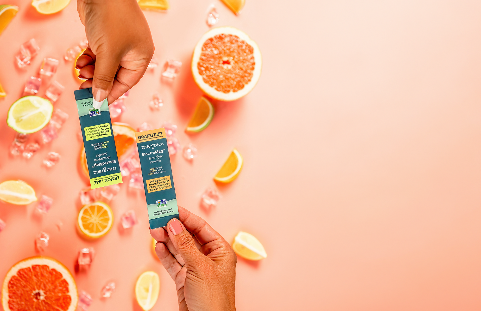 Two hands holding True Grace ElectroMag hydration drink mix packets with fruit slices on a pink background