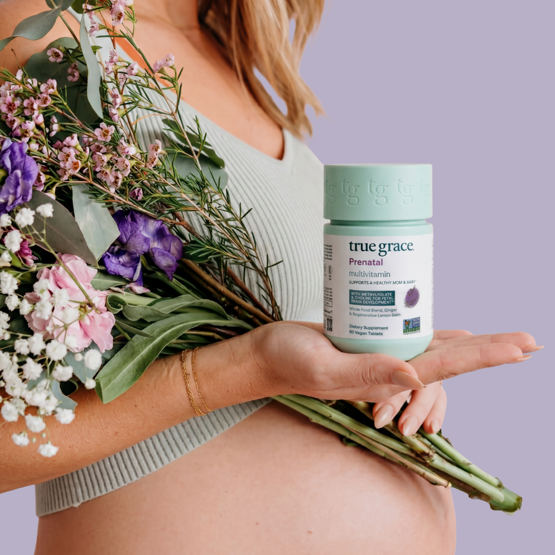 Pregnant person holding a bouquet of flowers and a container of True Grace prenatal vitamins against a purple background.