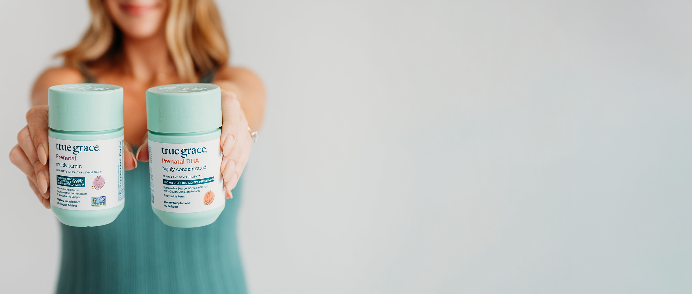 Woman holding bottles of True Grace Prenatal and Prenatal DHA vitamins against a plain background